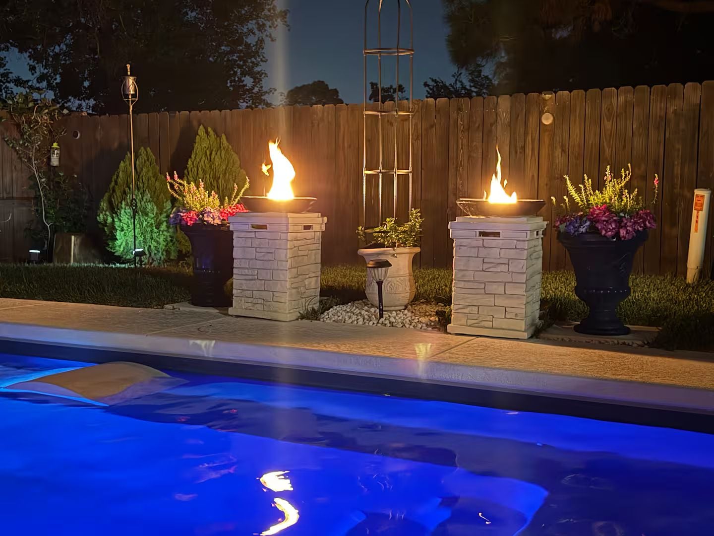 A pool area with three lit fire features, surrounded by a wooden fence and potted plants.
