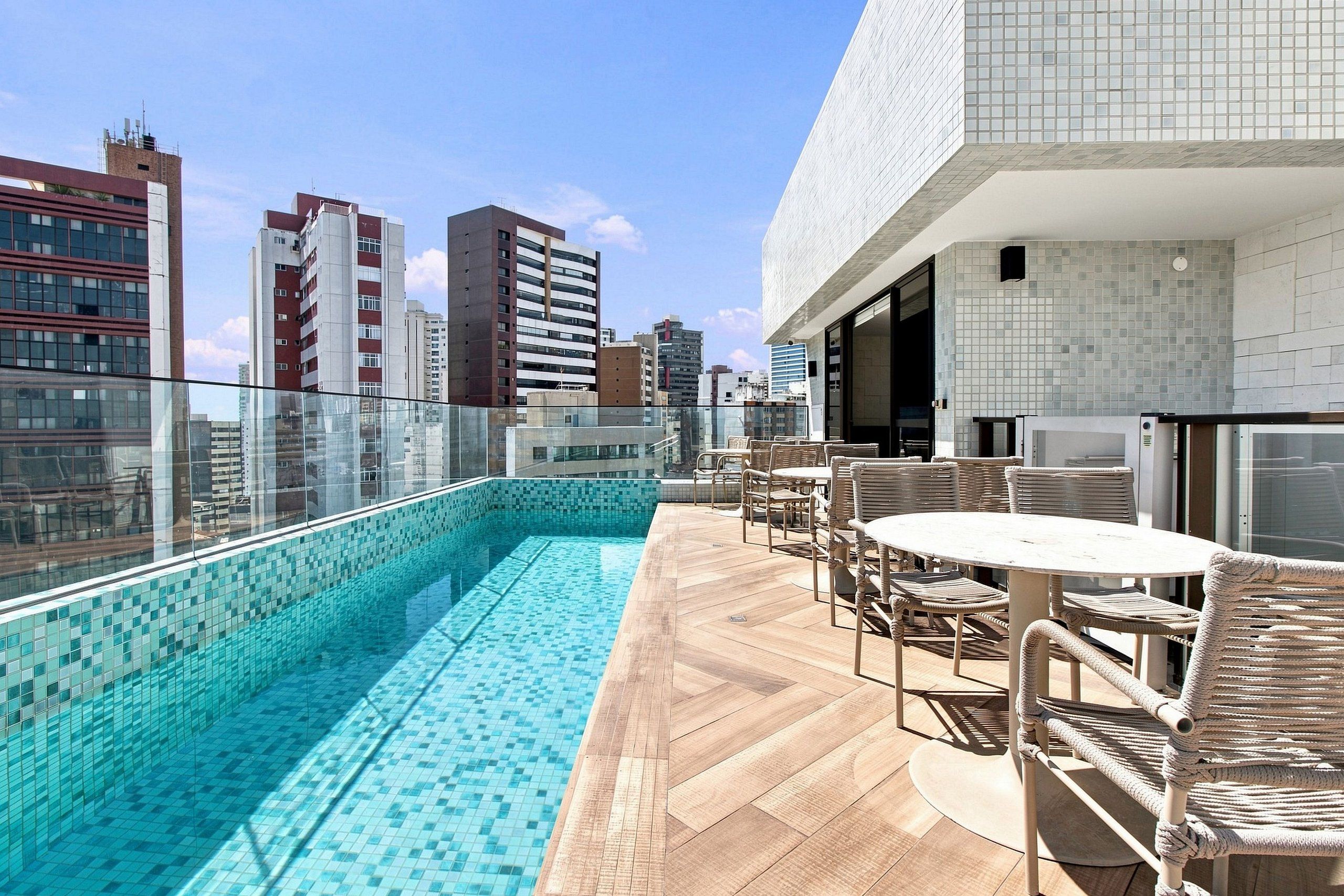 A rooftop pool area with a glass balustrade, wicker furniture, and a view of the city skyline.