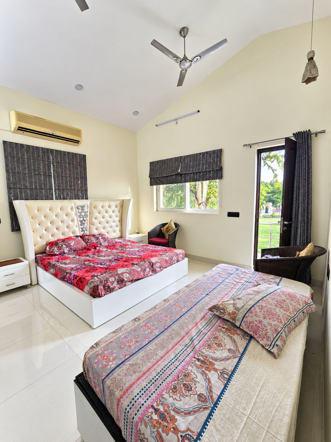 A bedroom with a large bed, a ceiling fan, an air conditioning unit, a window with curtains, and a view of greenery outside.