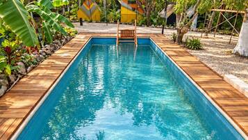 Una piscina rectangular con una terraza de madera alrededor, ubicada en un jardín tropical con diversas plantas y árboles.