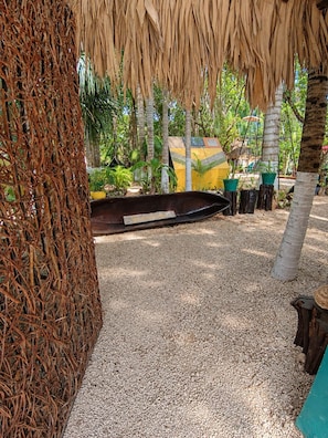 A tropical outdoor area with a thatched roof, gravel ground, and various potted plants.