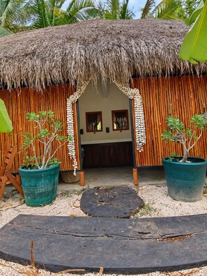 A thatched-roof hut with a wooden facade, two potted plants, and a wooden bench in the foreground.