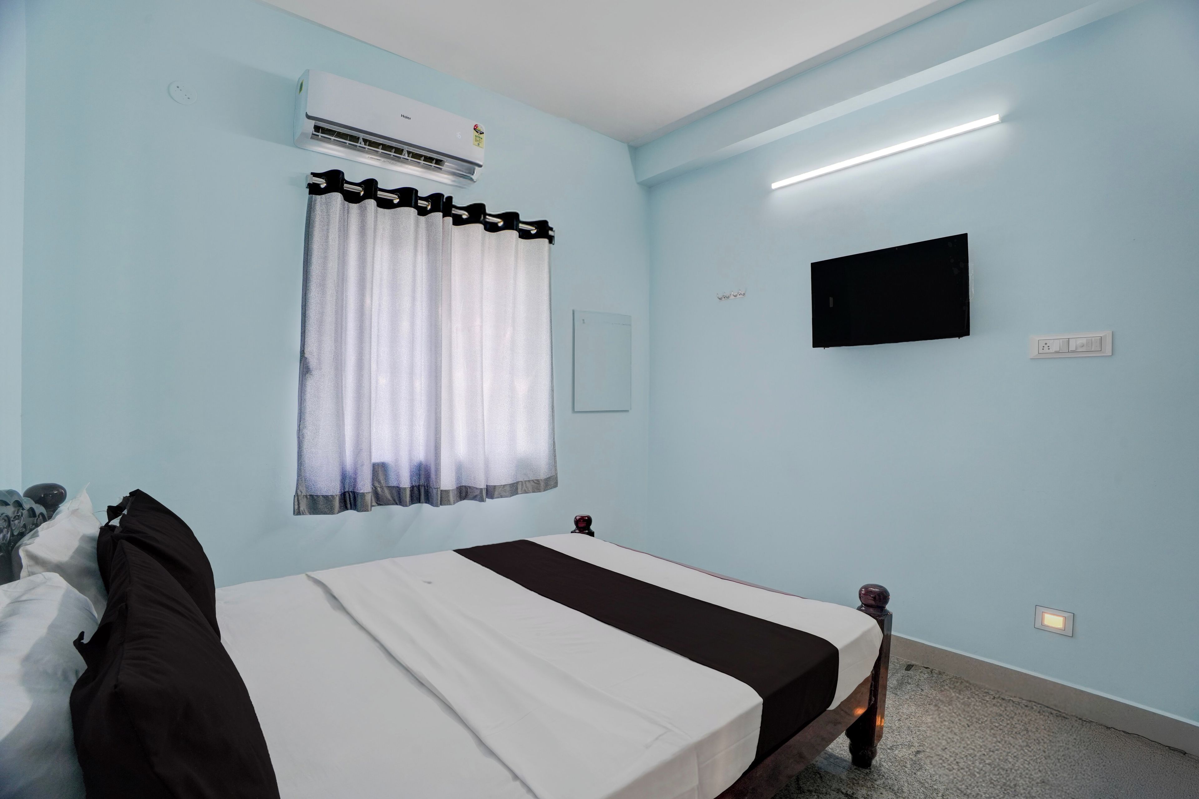 A hotel room with a bed, a window with curtains, an air conditioner, a flat-screen TV, and a wall socket.