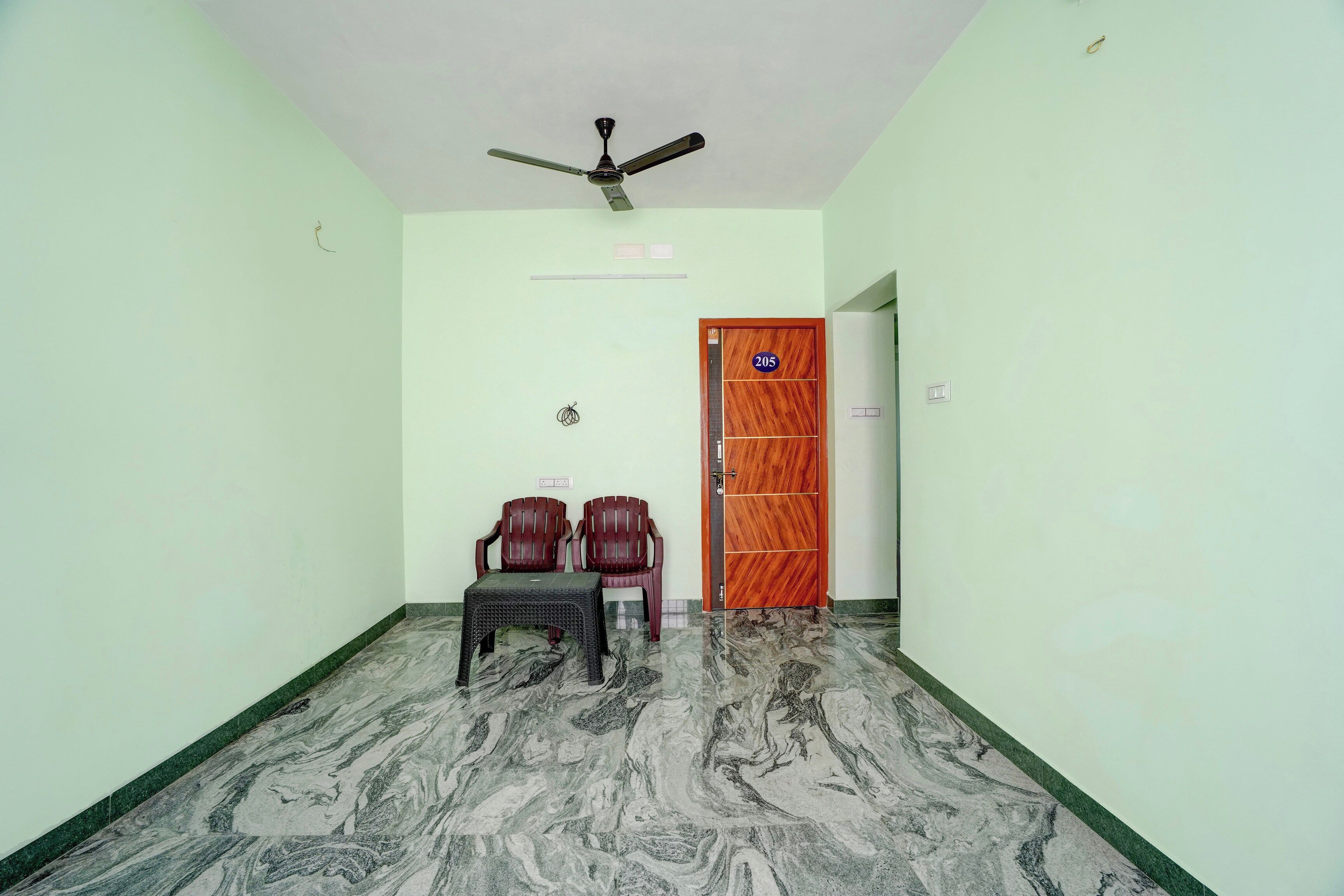 A room with a ceiling fan, a wooden door with a number plate, two plastic chairs, and a small table.