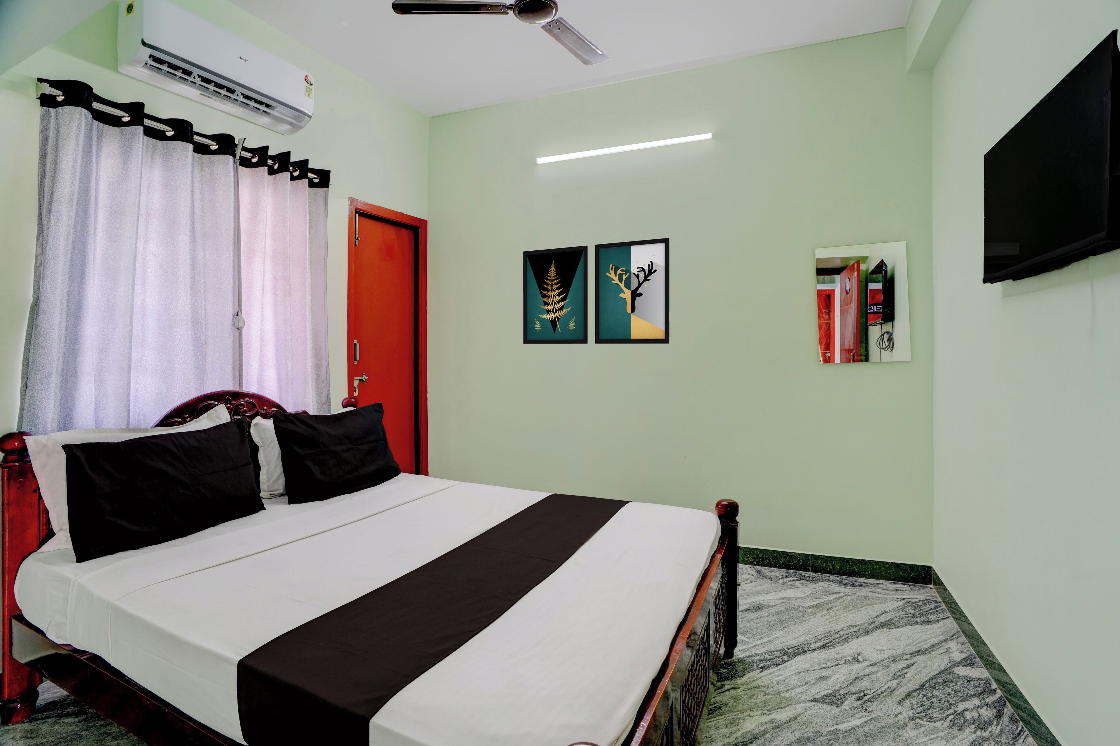 A hotel room with a bed, a red door, a ceiling fan, an air conditioner, and a flat-screen TV mounted on the wall.