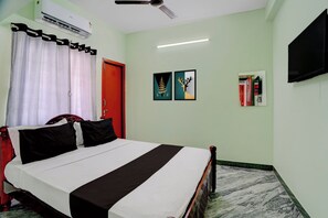 A hotel room with a bed, a red door, a ceiling fan, an air conditioner, and a flat-screen TV mounted on the wall.