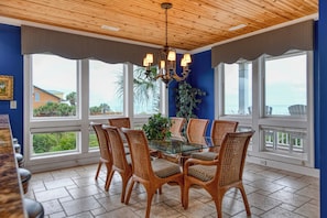 A dining area with a glass table surrounded by wicker chairs, large windows with view, and a wooden ceiling. - The Oasis - Seven Bedroom Oceanfront Home with a Private Pool and Hot Tub (Myrtle Beach)