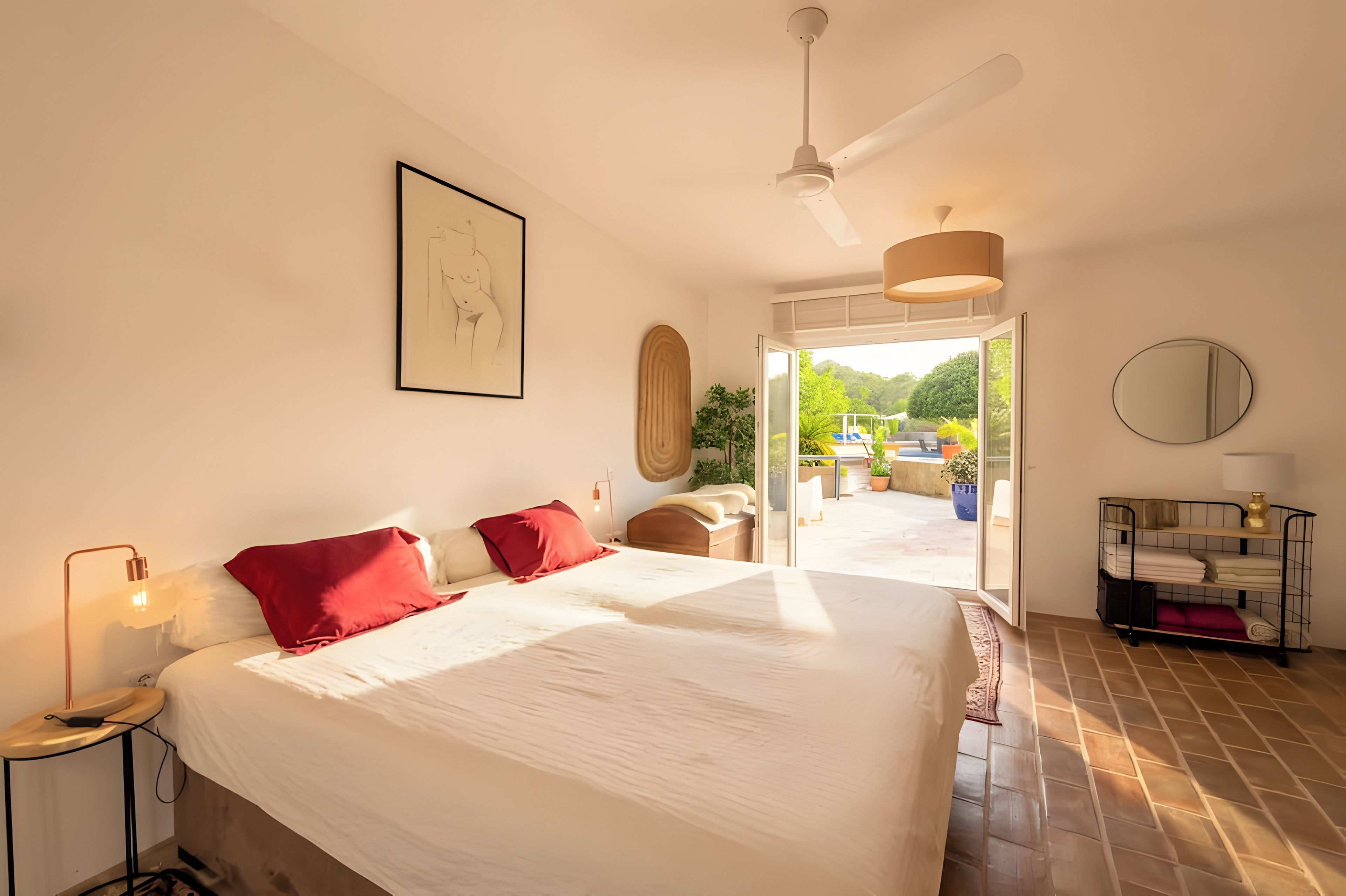 A hotel room with a large bed, a ceiling fan, a framed artwork, a side table with a lamp, and a view of the outdoors.