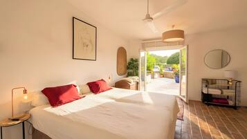 A hotel room with a large bed, a ceiling fan, a framed artwork, a side table with a lamp, and a view of the outdoors.