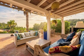 A covered outdoor seating area with a sofa, chairs, and a table, overlooking a landscaped garden and a body of water.