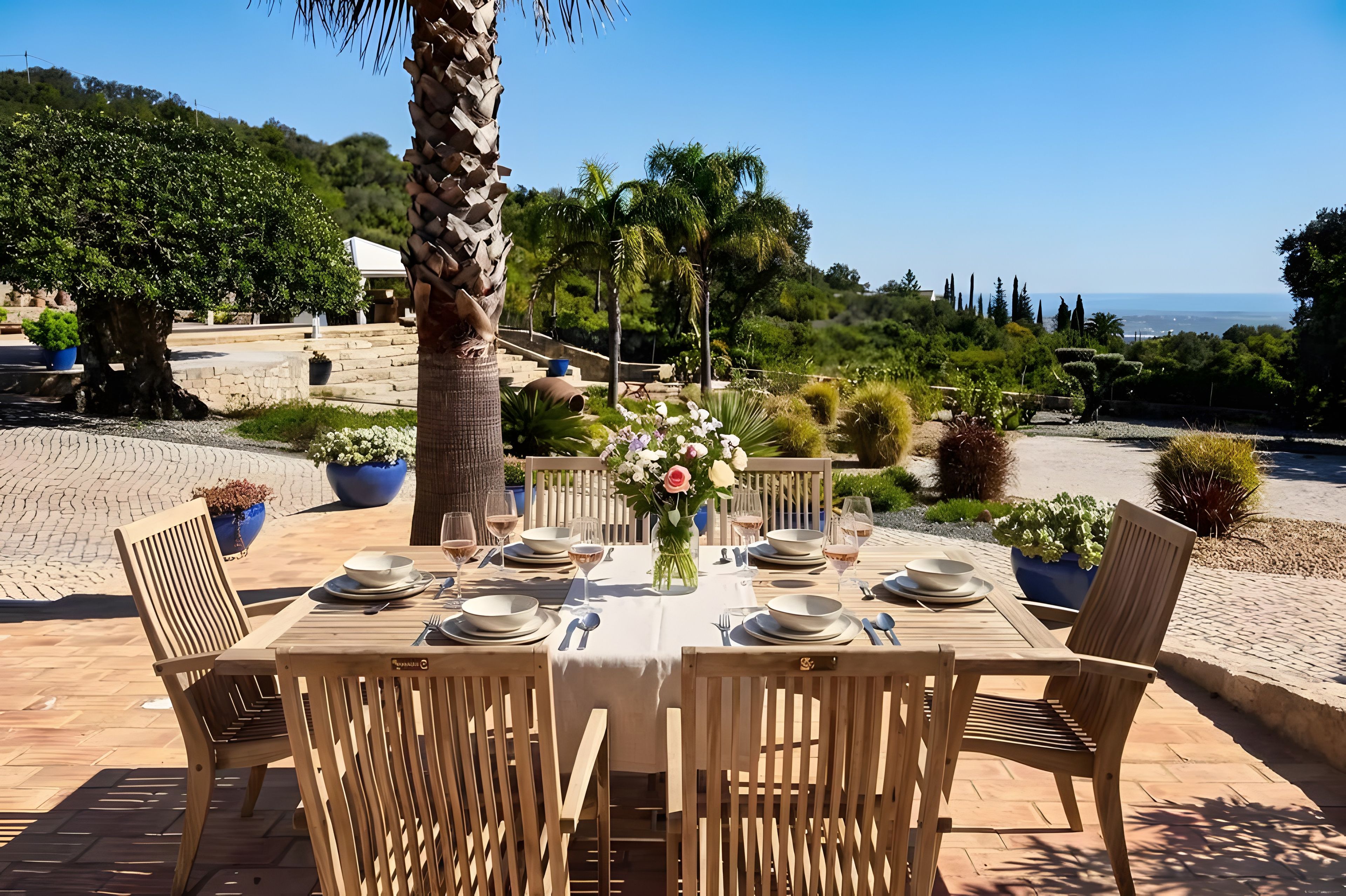 An outdoor dining area with a table set for four, surrounded by chairs, with a view of the sea and greenery in the background.