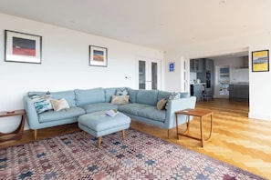 A living room with a light blue sectional sofa, a matching ottoman, and a patterned rug. - 5BR Beachfront Escape with Panoramic Sea Views (Middleton on Sea)