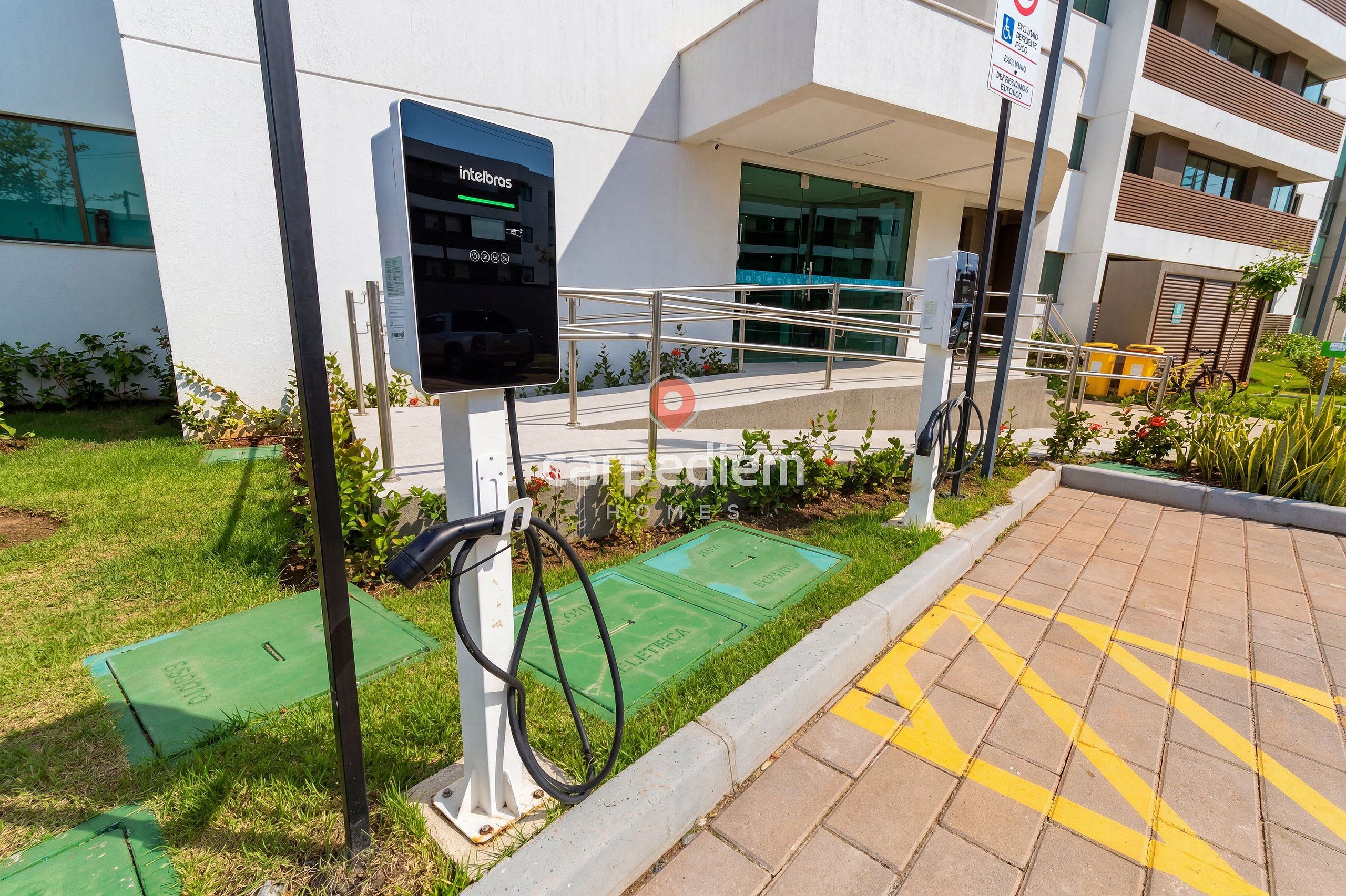 Una estación de carga para vehículos eléctricos al aire libre con pantalla digital, rampa accesible para sillas de ruedas y un edificio de diseño moderno al fondo.