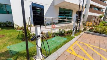 Una estación de carga para vehículos eléctricos al aire libre con pantalla digital, rampa accesible para sillas de ruedas y un edificio de diseño moderno al fondo.