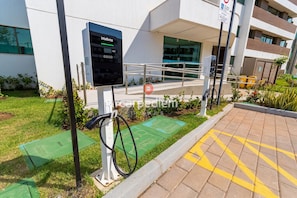 Uma estação de carregamento para veículos elétricos ao ar livre, com visor digital, rampa acessível para cadeiras de rodas e um edifício com design moderno ao fundo.