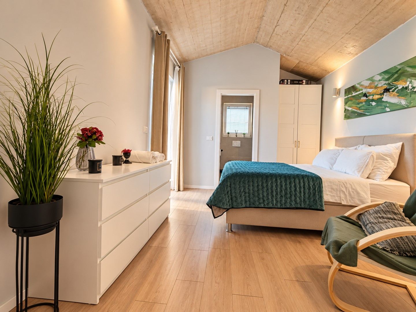 Ein Schlafzimmer mit einem Bett, einem Sessel, einem Nachttisch mit Pflanze und einem Bild an der Wand.