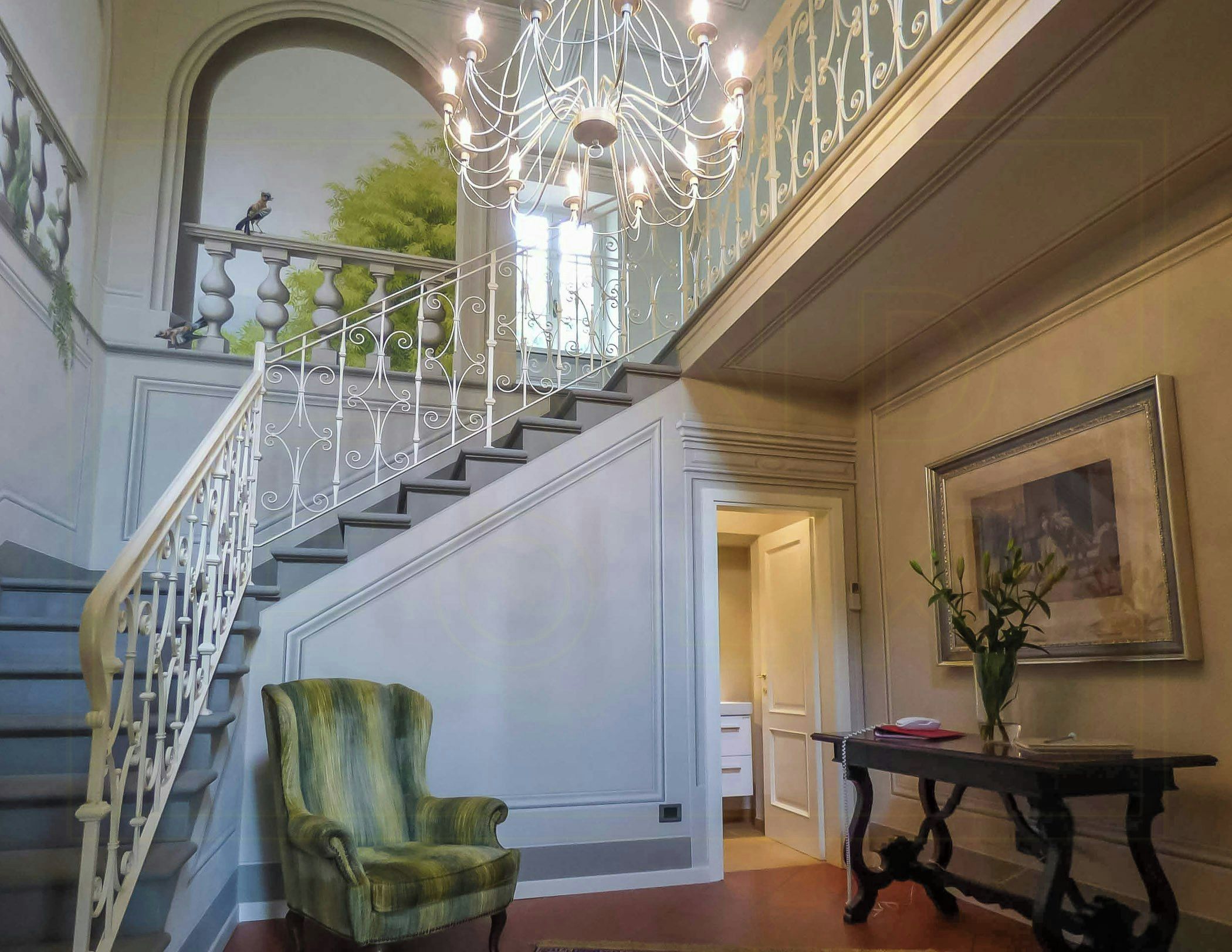 Una imponente escalera con barandales ornamentados, un candelabro y un sillón verde en un vestíbulo espacioso.