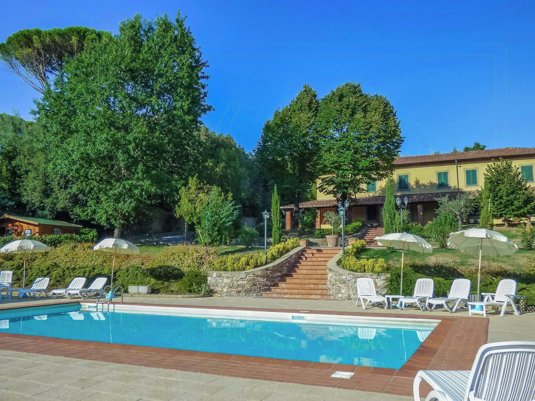 Una piscina con lettini e ombrelloni, circondata da alberi e un edificio con tetto in tegole.