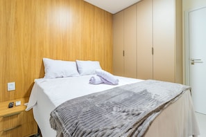 A hotel room with a wooden headboard, a bed with white linens, a grey blanket, and a bedside table with a remote control. - Cais Eco #311 - Cobertura Com Piscina by Carpediem (Ipojuca)