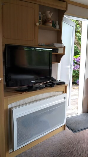 A television on a wooden stand with a white vent below it, a small shelf with decorative items, and a door leading to a garden.