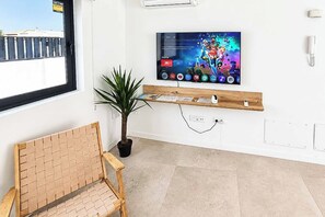 A room with a television mounted on the wall, a wooden shelf below it, a potted plant, and a brick-patterned chair. - Casa Drago Uno - Three Bedroom Villa, Sleeps 5 (Caleta de Fustes)