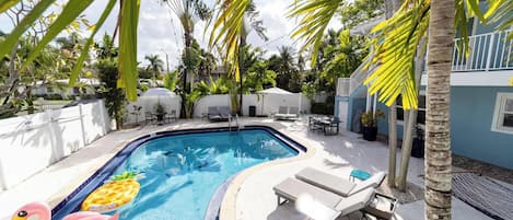 A swimming pool with lounge chairs and umbrellas, surrounded by palm trees and a white fence.