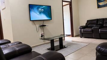 A living room with a flat-screen TV mounted on the wall, leather sofas, a glass coffee table, and a door leading to another room.