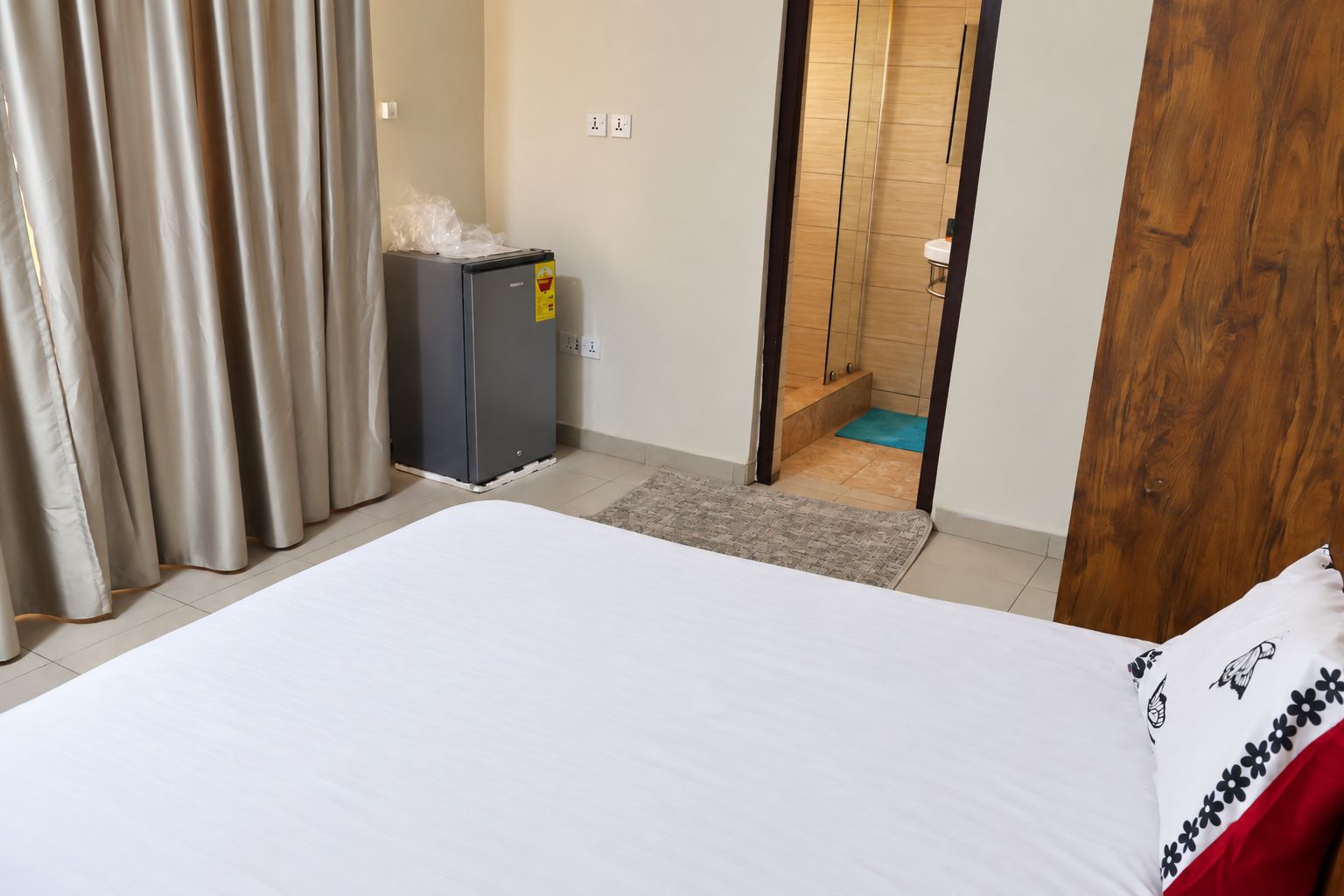 A hotel room with a bed, a small refrigerator, a bathroom visible through an open door, and a wooden door frame.