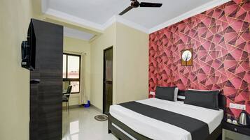 Habitación de hotel con cama, ventilador de techo, televisión y pared con un estampado geométrico rojo.