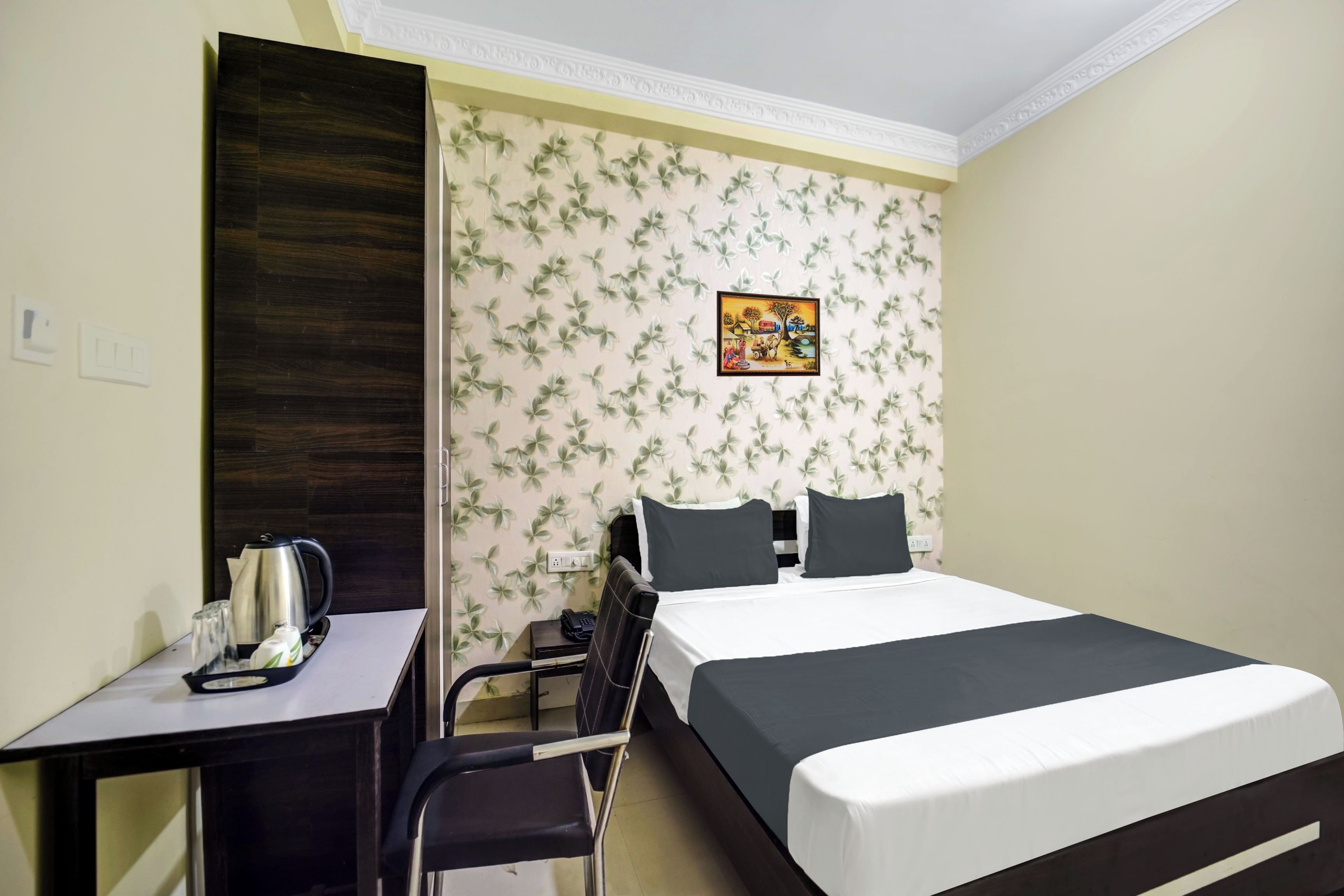 A hotel room with a bed, a desk with a kettle, a chair, and a wall with a floral pattern.