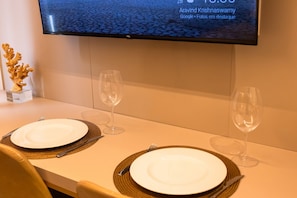 Una mesa de comedor elegantemente dispuesta con dos platos, cubiertos y copas de vino, un adorno de coral y un televisor que muestra una escena urbana.