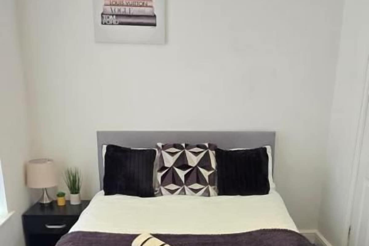 A neatly made bed with a dark-colored bedspread and two pillows, a small bedside table with a lamp, and a framed picture on the wall.