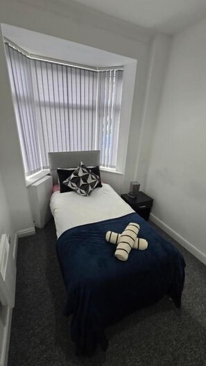 A small, neatly arranged bedroom with a single bed, a bedside table, a window with blinds, and a plush toy on the bed. - Single & Double rooms in Stoke on trent (Stoke-on-Trent)