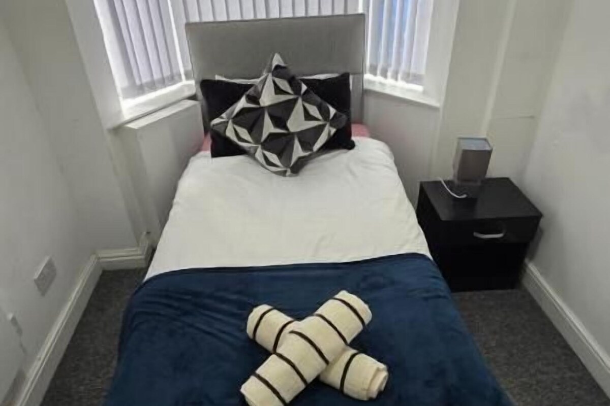 A small, neatly arranged bedroom with a bed, a nightstand, and a window with blinds.
