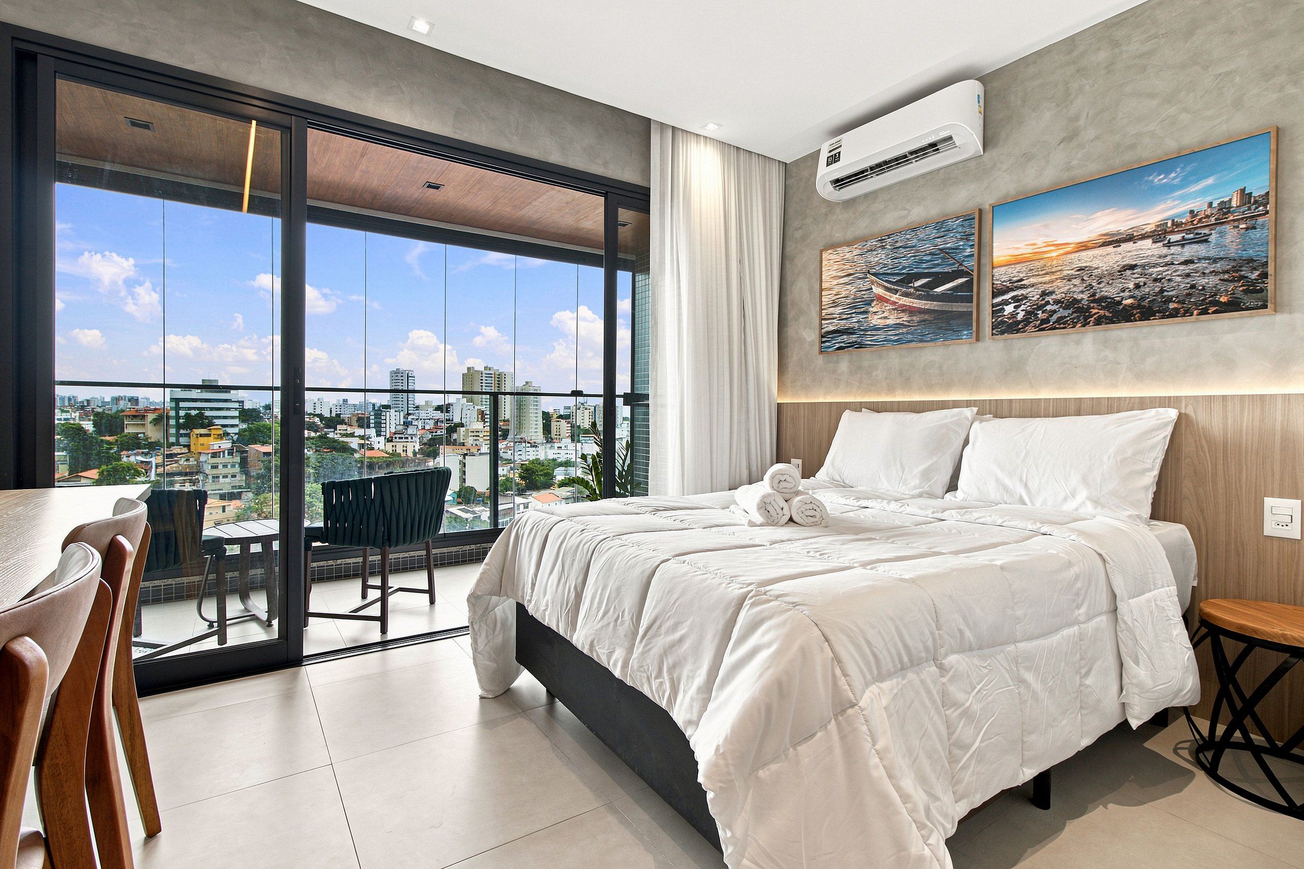 A modern hotel room with a large balcony, a bed with white linens, and two cityscape paintings on the wall.