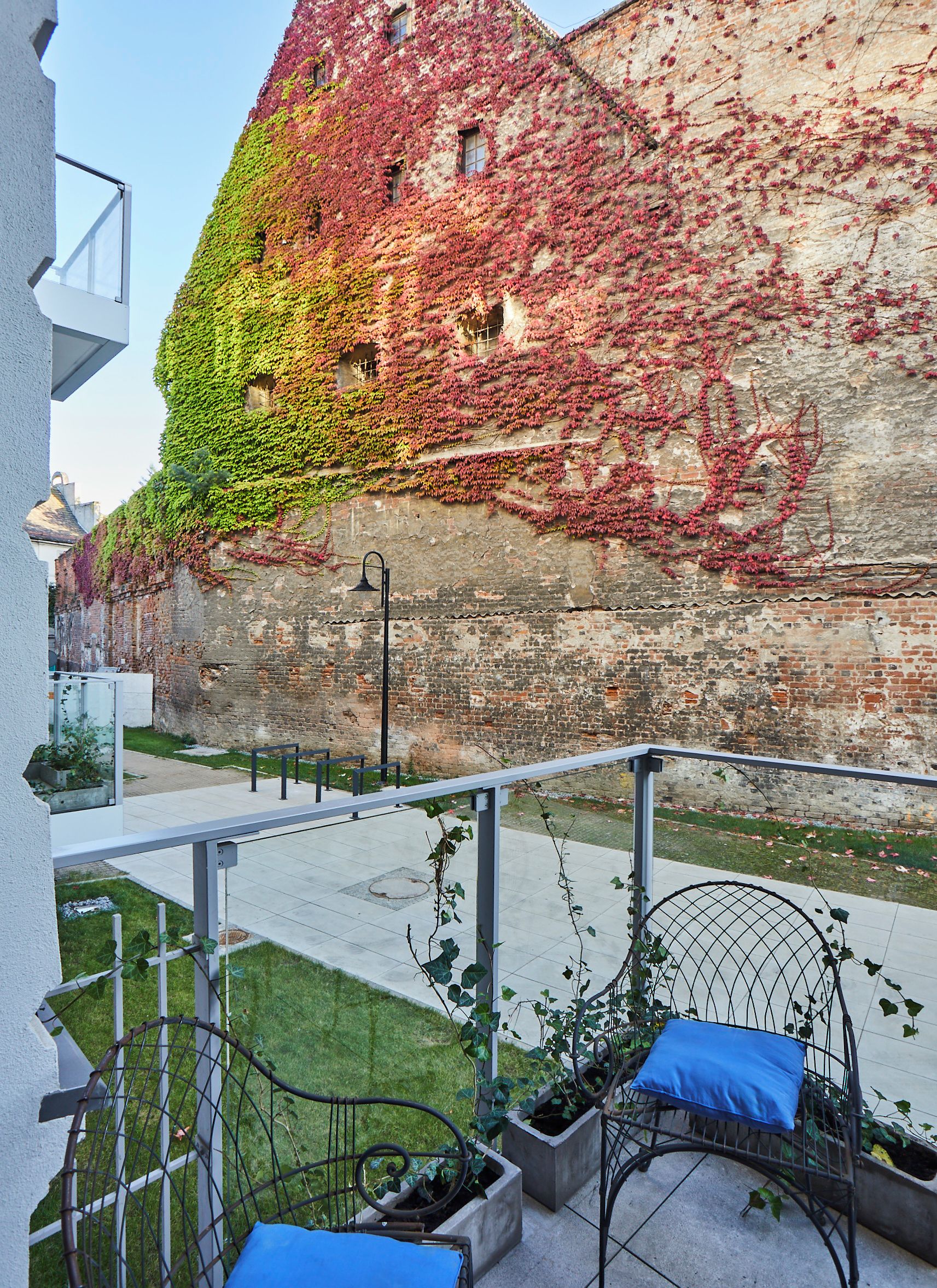 Une cour avec un mur de briques recouvert de lierre, un banc en métal avec un coussin et un portail en métal.