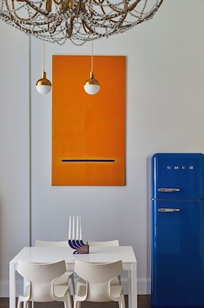 Ein Essbereich mit einem weißen Tisch und Stühlen, einem blauen Kühlschrank und einem orangefarbenen abstrakten Gemälde an der Wand.