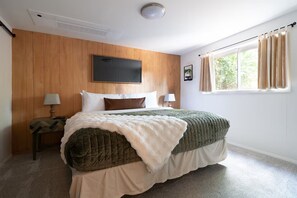 Ein Schlafzimmer mit einem Bett, zwei Nachttischen, einem fensterförmigen Öffnung mit Vorhängen und einem an der Wand befestigten Fernseher.