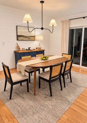 Een eetruimte met een houten tafel, vier stoelen, een kroonluchter, een schilderij aan de muur en uitzicht door een glazen deur.