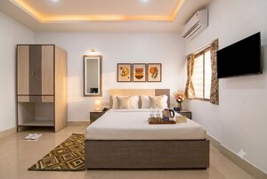 A hotel room with a bed, bedside table, lamp, artwork, wardrobe, and a TV.