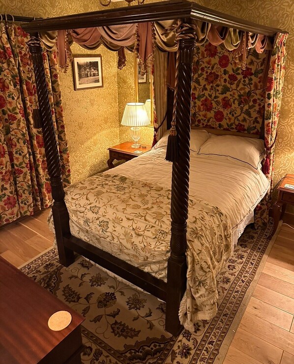 Ein Himmelbett mit Blumenmuster, ein Nachttisch mit Lampe und ein gemustertes Teppich.