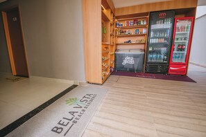 A hotel lobby with a reception desk, a refrigerator stocked with beverages, and a wooden shelving unit with snacks. - MG01J  Bella V.  Ap. 105 B  Andres Sardi (Ipojuca)