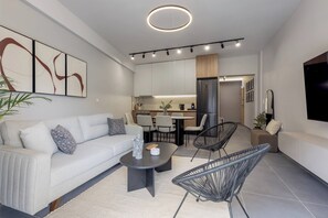 A modern living room with a sofa, coffee table, and chairs, a kitchen area with a dining table, and a TV mounted on the wall. - Elegant Urban Flat Near Center (Kallithea)