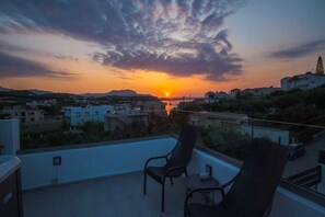 A balcony with two chairs and a table, overlooking a sunset with a cityscape in the background. - Sea-View Villa with Rooftop Hot Tub (Crete)