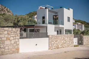 A modern two-story house with a stone wall, a balcony, and a view of the surrounding landscape. - Sea-View Villa with Rooftop Hot Tub (Crete)