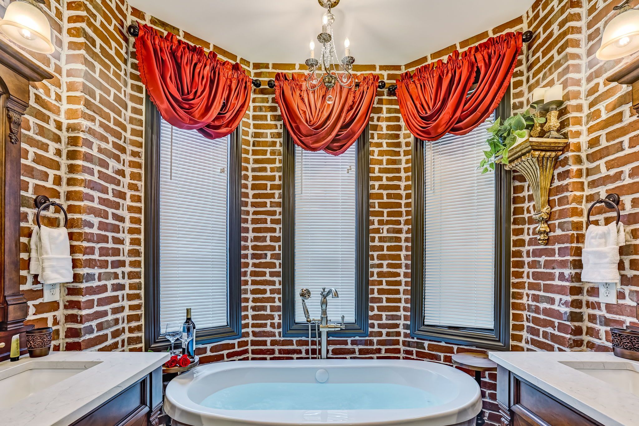 A bathroom with a large bathtub, three windows with red curtains, and a chandelier.