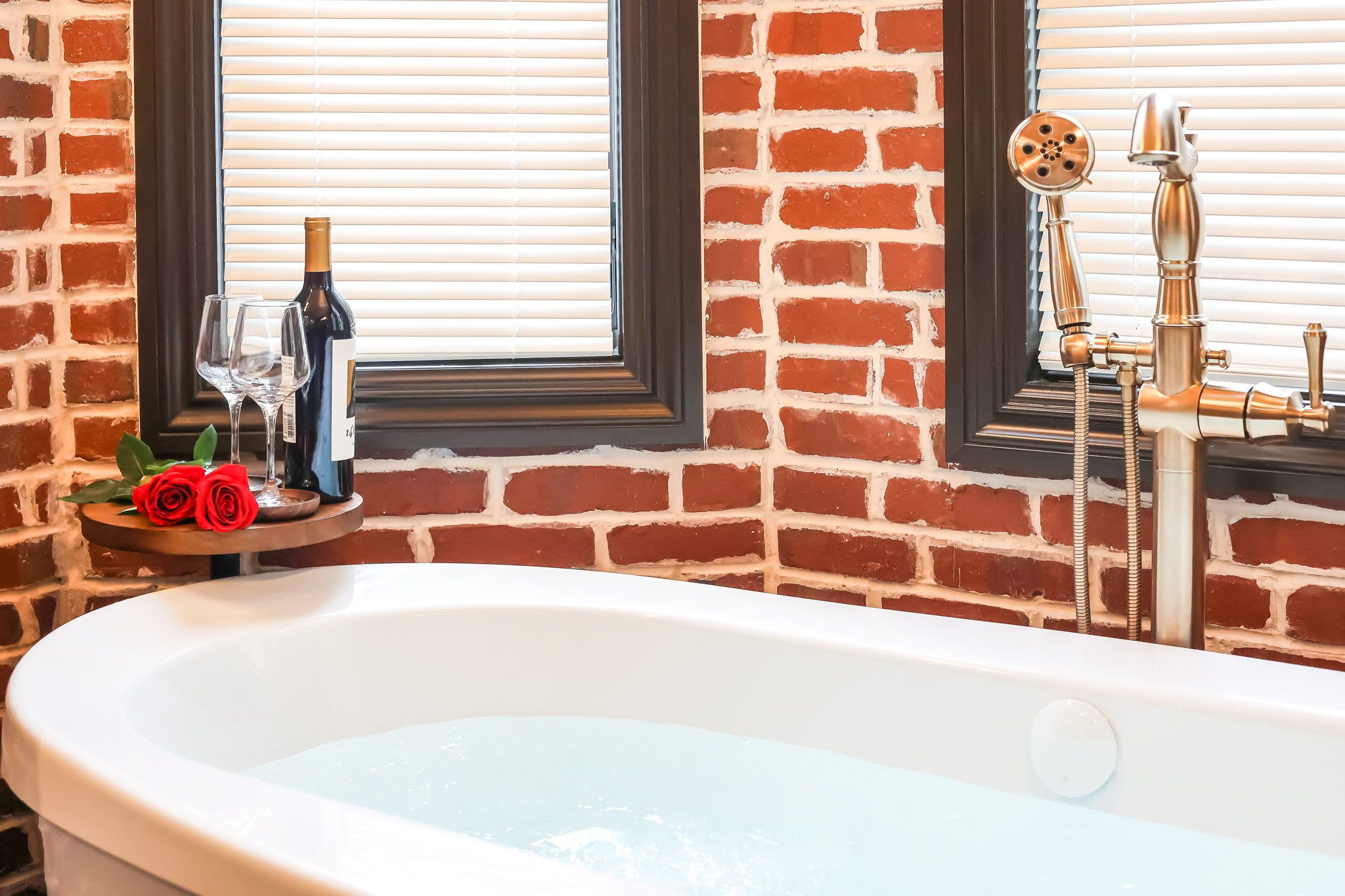 A bathroom with a white bathtub, a window with blinds, a brick wall, and a mirror with a faucet.