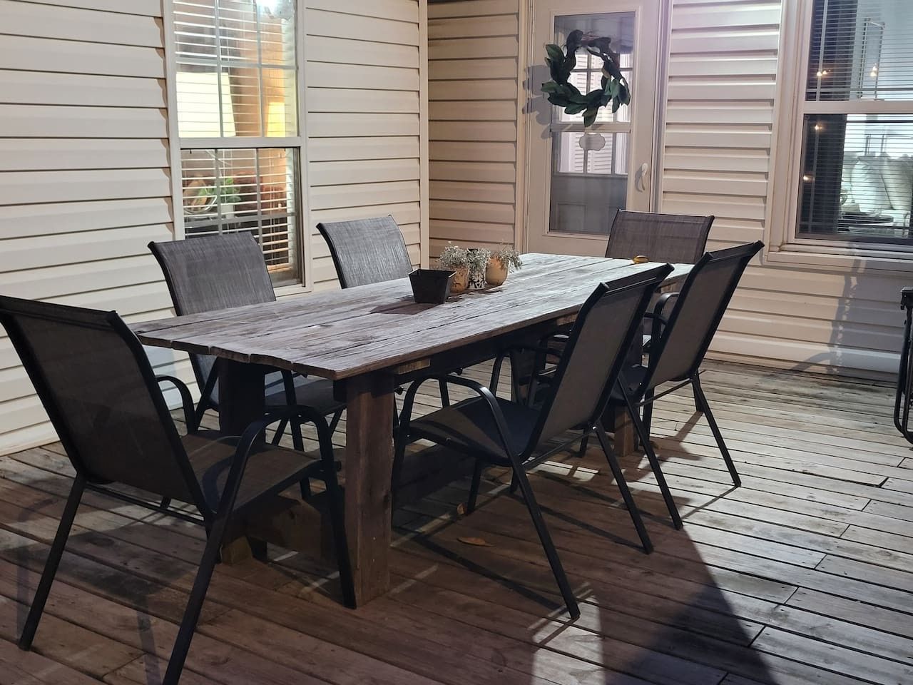 An outdoor patio with a wooden table and chairs, a potted plant, and a window with blinds.