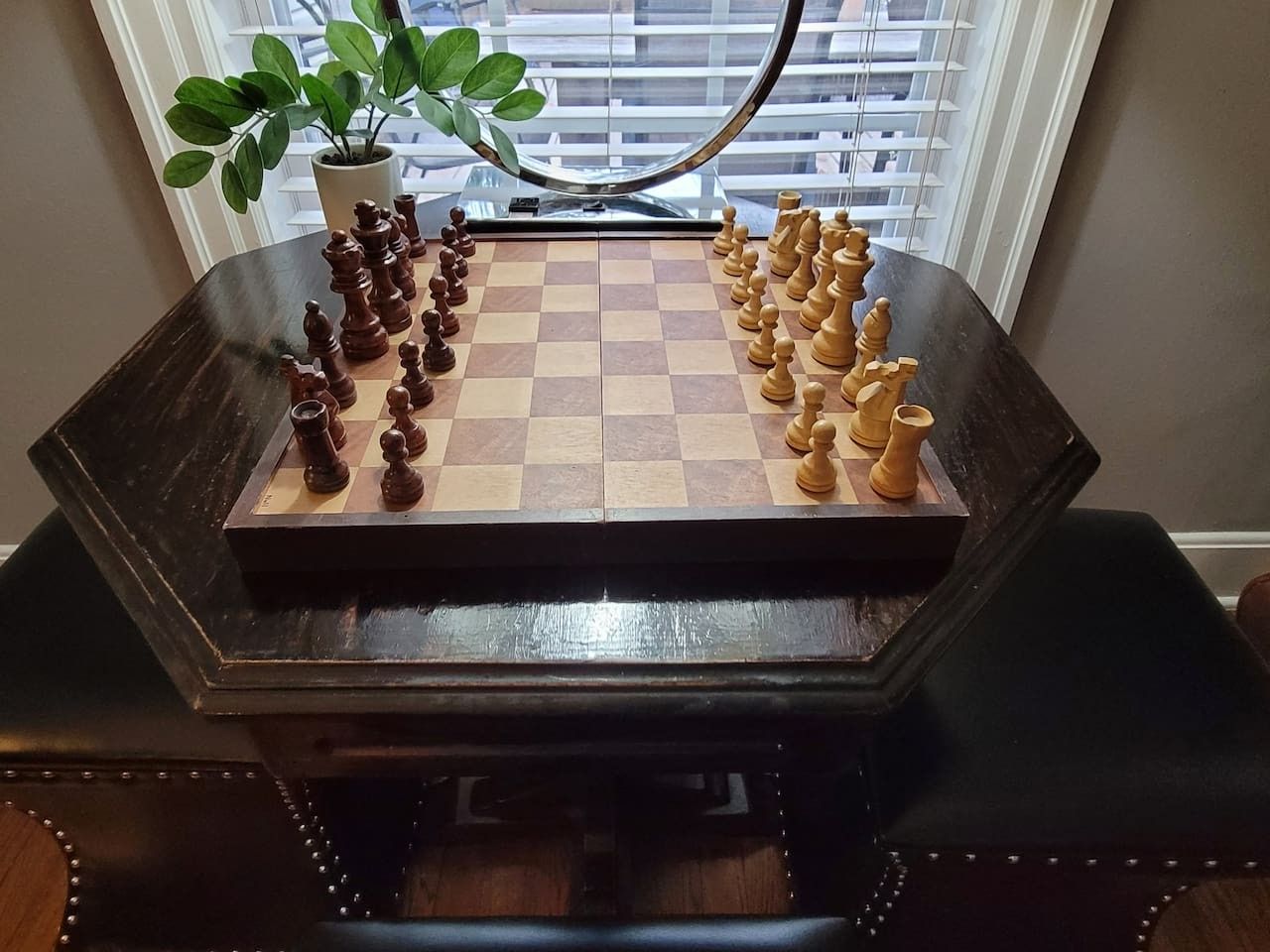 A chessboard set up on a dark wooden table with a potted plant beside it.
