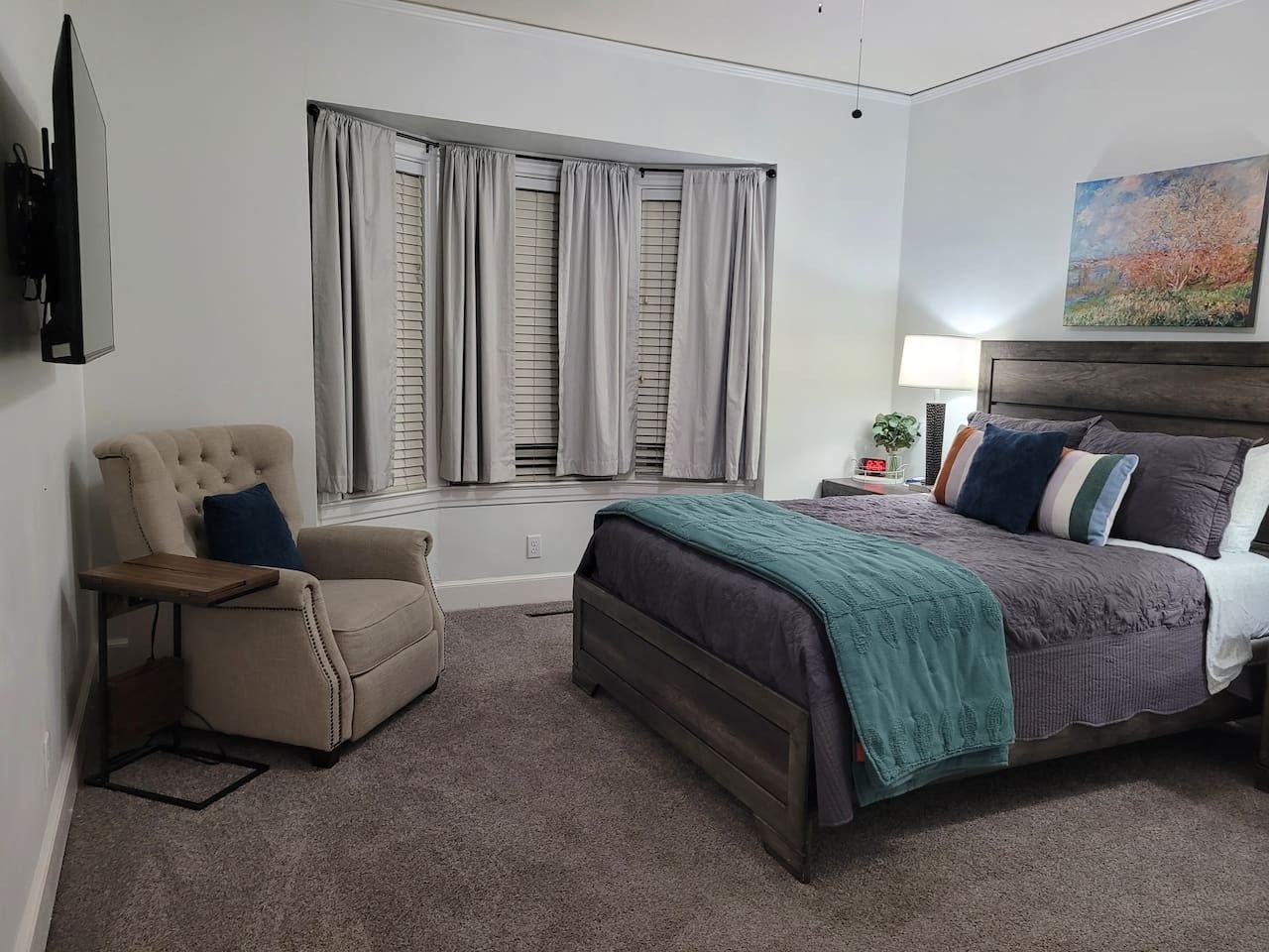 A bedroom with a bed, a chair, a nightstand, a lamp, and a window with curtains.